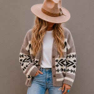 Peut inclure: Un chapeau en feutre beige à larges bords avec une bande brune texturée et une plume, porté avec un gilet boutonné à motif géométrique noir et blanc sur fond beige, un t-shirt blanc uni et un jean en denim bleu clair.