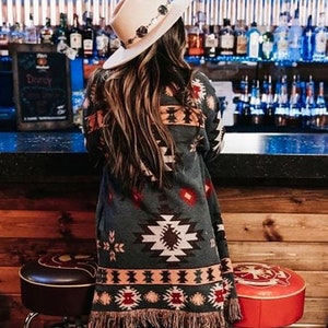 May include: A woman wearing a cream-coloured cowboy hat with decorative accents and a patterned, fringed cardigan. The cardigan features a geometric design in shades of grey, red, and white. The background shows a bar with bottles and stools.