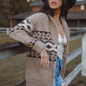 Peut inclure: Un long cardigan ouvert sur le devant, dans une palette de couleurs neutres, avec un motif géométrique noir, blanc et vert. Le cardigan a un col châle et est porté avec un haut blanc et un jean bleu. Un chapeau noir et un collier complètent le look.