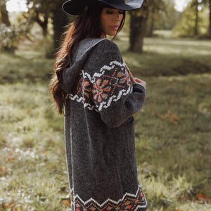 May include: A dark grey hooded cardigan with a patterned design in shades of orange, pink, and white. The long-sleeved sweater has a black hat. The cardigan is knee-length and appears to be made of a soft, knitted material.