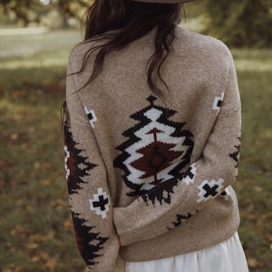 May include: A tan sweater with a geometric pattern in black, white, and brown is worn with a brown cowboy hat. The sweater has a detailed design on the back and sleeves. The person is standing outdoors in a natural setting.