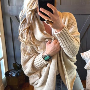 May include: A beige open-front knit cardigan featuring a textured waffle-knit pattern on the body, wide ribbed cuffs, and a draped shawl collar. The garment is worn over light blue denim jeans with a distressed knee detail.