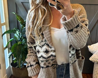 May include: A beige and black patterned cardigan with a shawl collar and button closure. The long-sleeved sweater has pockets and a geometric design on the front and sleeves. The cardigan is paired with a white top and blue jeans.