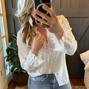 May include: A white long-sleeved blouse with detailed lace cuffs and bodice. The blouse has a button-down front and a V-neck. The model is wearing light blue denim jeans.