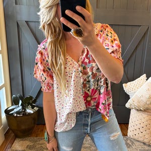 Könnte beinhalten: Ein Patchwork-Top mit V-Ausschnitt und kurzen Ärmeln. Das Oberteil hat eine Mischung aus Blumen-, geometrischen und gepunkteten Mustern in Rosa, Weiß und anderen Farben. Das Model trägt eine hellblaue Jeans.