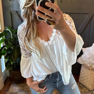 Puede incluir: Top blanco de crochet con mangas acampanadas y ribete de encaje. El top se lleva con jeans azul claro.