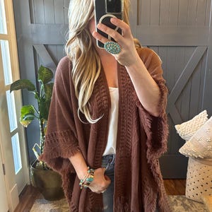 Puede incluir: Un poncho de punto marrón con detalles de flecos. El poncho tiene un corte holgado y está abierto por delante. La modelo lleva el poncho sobre un top blanco de tirantes y unos vaqueros azules.