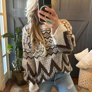 Puede incluir: Un suéter de color marrón y blanco con cuello alto. El suéter tiene un diseño geométrico con colores marrón, blanco y crema.