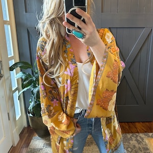 May include: A long, yellow floral kimono with pink and brown accents. The kimono has a wide, flowing silhouette and a tie closure at the waist.
