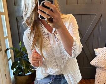 May include: A white lace blouse with a button-up front and 3/4 sleeves. The blouse is worn over a pair of light blue jeans.