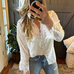 May include: White long-sleeve blouse with intricate lace detailing on the sleeves and bodice. The blouse features a button-down front and a textured fabric. The model is wearing light-wash blue jeans.