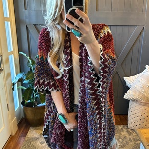 May include: A maroon, blue, and white patterned cardigan with fringe trim. The cardigan has a geometric design and is open in the front. The model is wearing the cardigan over a white tank top and blue jeans.