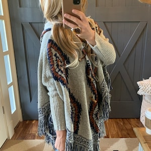 Peut inclure: Un poncho à capuche gris avec un motif géométrique dans les tons de brun, bleu et blanc. Le poncho est doté d'une bordure à franges et d'une fermeture à bouton.