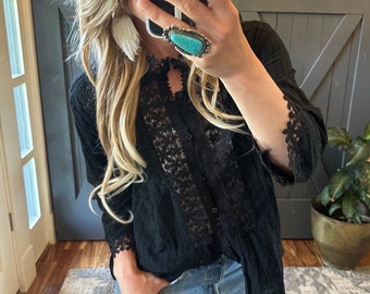 May include: A black lace blouse with a button-up front and long sleeves. The blouse has a sheer lace overlay and a delicate lace trim around the cuffs and neckline. The model is wearing blue jeans and a turquoise ring.