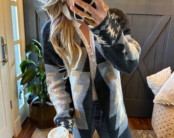 May include: A gray and beige cardigan with a geometric pattern. The cardigan is open in the front and has long sleeves. The person is wearing blue jeans and a white shirt.