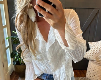May include: White long-sleeve blouse with a ruffled neckline and bell sleeves. The blouse has a dotted pattern and is made of a sheer fabric.