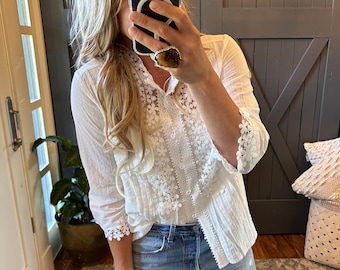 May include: A white, semi-sheer blouse with a lace overlay and floral embroidery. The blouse has a button-up front and 3/4 sleeves.