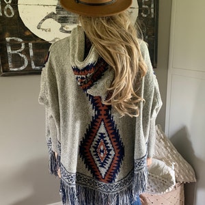 Op de afbeelding: Een grijze poncho met capuchon met een geometrisch patroon in tinten bruin, blauw en wit. De poncho heeft franjes langs de onderkant en is gemaakt van een zachte, gebreide stof.