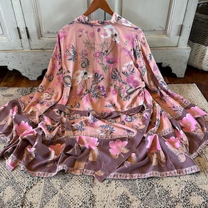 May include: A peach-colored floral print kimono-style robe with a brown border. The robe features a variety of pink and purple flowers and foliage. The robe is hanging on a wooden hanger.