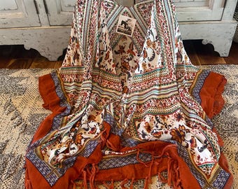 Op de afbeelding: Een kimono-achtig kledingstuk met een crèmekleurige achtergrond en een bloemenpatroon in oranje, rood en groen. De randen zijn afgewerkt met een effen roestkleurige stof en franjes.