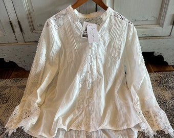 May include: Cream-colored blouse with long sleeves and intricate lace detailing. The blouse features a high collar, button-down front, and delicate embroidery. The sleeves have flared cuffs with lace trim. A tag with the text "lavender & fig" is attached.