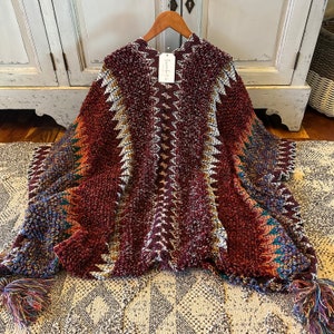 May include: A burgundy poncho with a textured, raised pattern and a zig-zag design in white, gold, and teal. The poncho has a hood and tassels at the ends. The tag reads "Emmalyn Taylor".