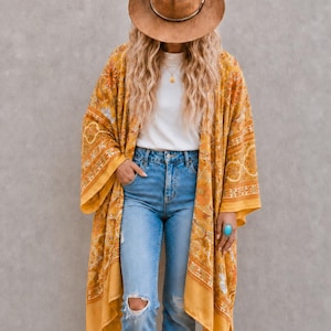 May include: A brown felt fedora hat, a white short-sleeved t-shirt, and a long, open-front mustard yellow kimono with a white floral pattern and tassel trim. The outfit includes light blue denim jeans with distressed knees and a turquoise ring.