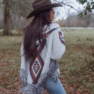May include: A woman wearing a brown cowboy hat and a grey poncho with a geometric pattern in shades of brown, navy, and white. The poncho has fringe along the bottom. She is wearing blue jeans.
