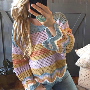Puede incluir: Un suéter de crochet a rayas de colores pastel en tonos azules, morados, naranjas y amarillos. El suéter presenta mangas acampanadas y un dobladillo festoneado. También se ve un anillo turquesa.