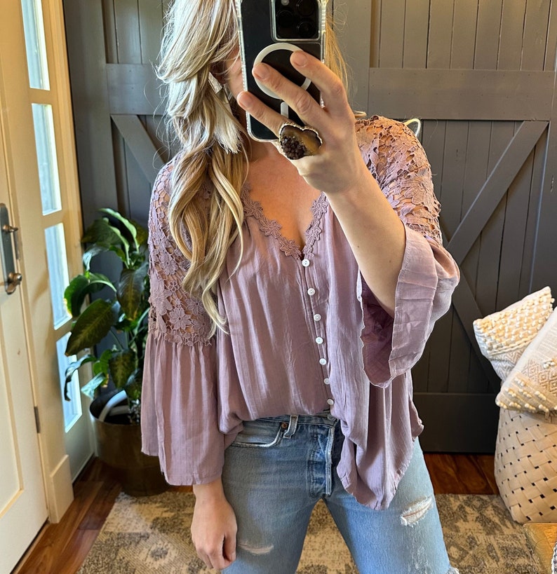 May include: A mauve button-up blouse with a lace overlay on the sleeves. The blouse has a relaxed fit and a v-neckline.