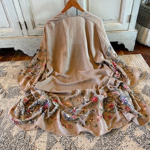 May include: A beige kimono-style robe featuring long, wide sleeves and a flowing hemline. The garment is decorated with intricate floral embroidery in shades of red, blue, green, and yellow along the sleeve edges and the bottom border.