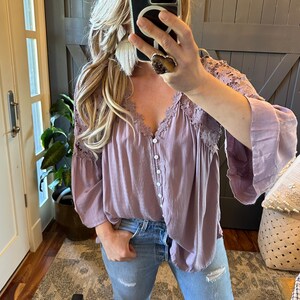 May include: A mauve button-up blouse with lace detailing on the neckline and sleeves. The blouse is made of a lightweight fabric and has a relaxed fit.