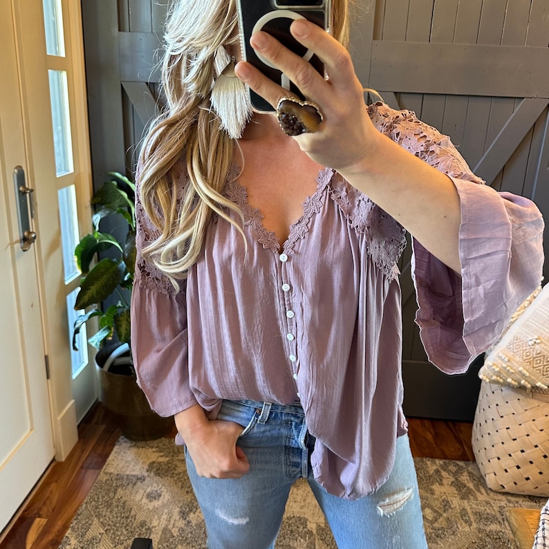 May include: A mauve button-up blouse with lace detailing on the neckline and sleeves. The blouse is made of a lightweight fabric and has a relaxed fit.