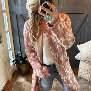 May include: A long, open-front cardigan in a dusty rose color with a cream-colored floral pattern. The cardigan has long sleeves and two front pockets. The interior lining is a light tan color. The model is wearing a white top and blue jeans.