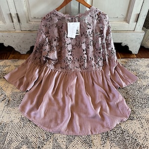 May include: A mauve-colored, lightweight, flowy top with a lace overlay. The lace is a delicate floral pattern. The top has a ruffled hem and 3/4 sleeves.