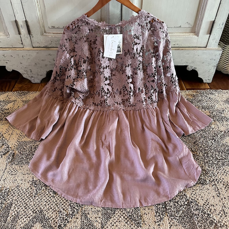May include: A mauve-colored, lightweight, flowy top with a lace overlay. The lace is a delicate floral pattern. The top has a ruffled hem and 3/4 sleeves.