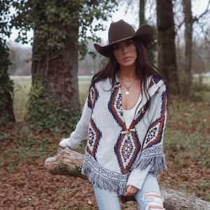 May include: A woman wearing a brown cowboy hat and a gray poncho with a geometric pattern in navy blue, rust, and white. The poncho has fringe along the bottom. She is wearing a white top and blue jeans.