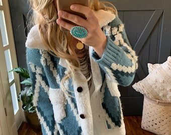 May include: A blue and white patterned cardigan with a white sherpa collar and a button closure. The cardigan has a relaxed fit and is perfect for layering.