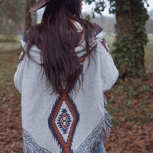 May include: A gray hooded poncho with a geometric pattern in shades of orange, navy, and white. The poncho has a fringed hem and is worn with a brown cowboy hat. The overall style is bohemian and casual.