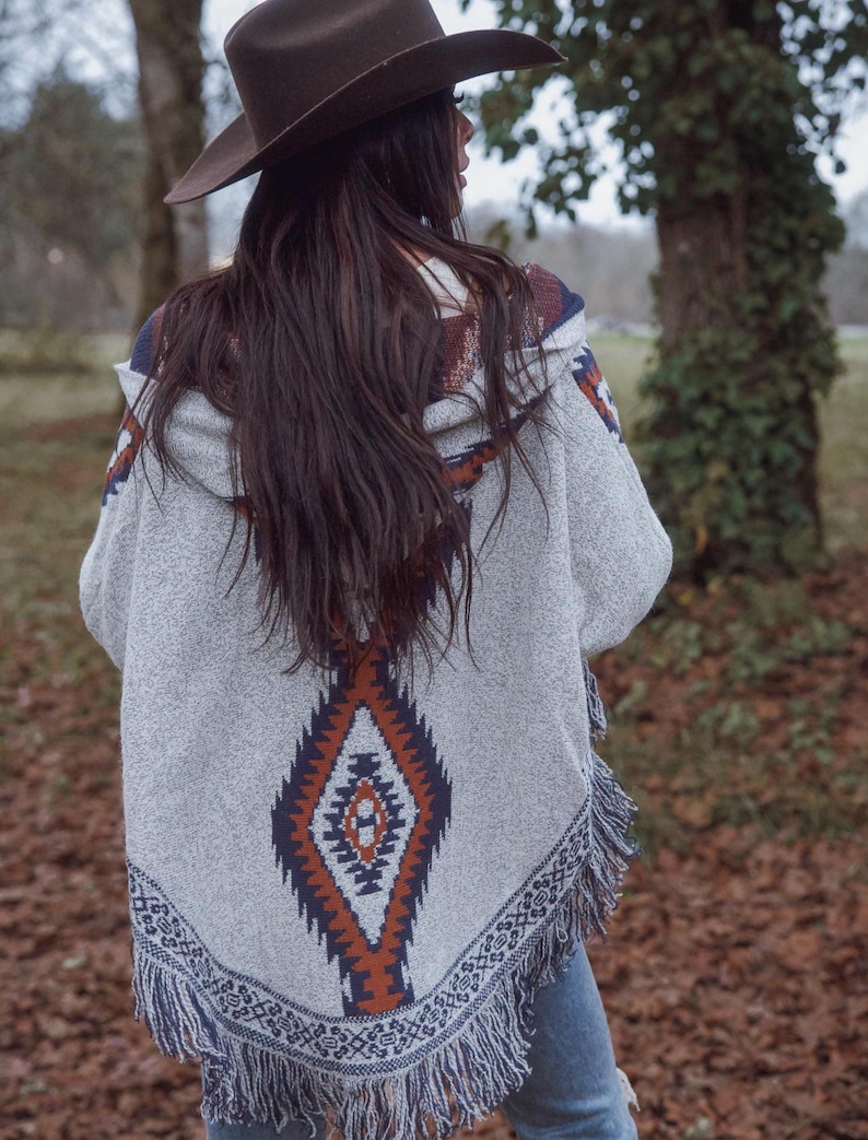 May include: A gray hooded poncho with a geometric pattern in shades of orange, navy, and white. The poncho has a fringed hem and is worn with a brown cowboy hat. The overall style is bohemian and casual.