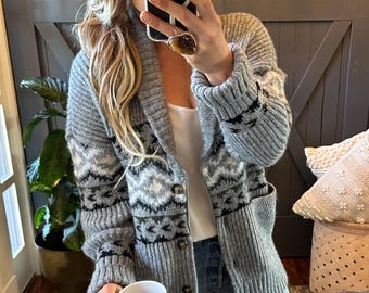 May include: A grey knit cardigan featuring a shawl collar, long sleeves, and a repeating geometric pattern in black and white. The garment has a button-front closure and two front pockets, worn over a white top and blue denim jeans.