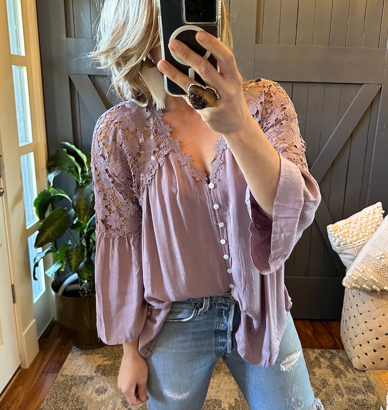 May include: A mauve button-up blouse with a lace overlay on the sleeves. The blouse has a relaxed fit and a v-neckline.