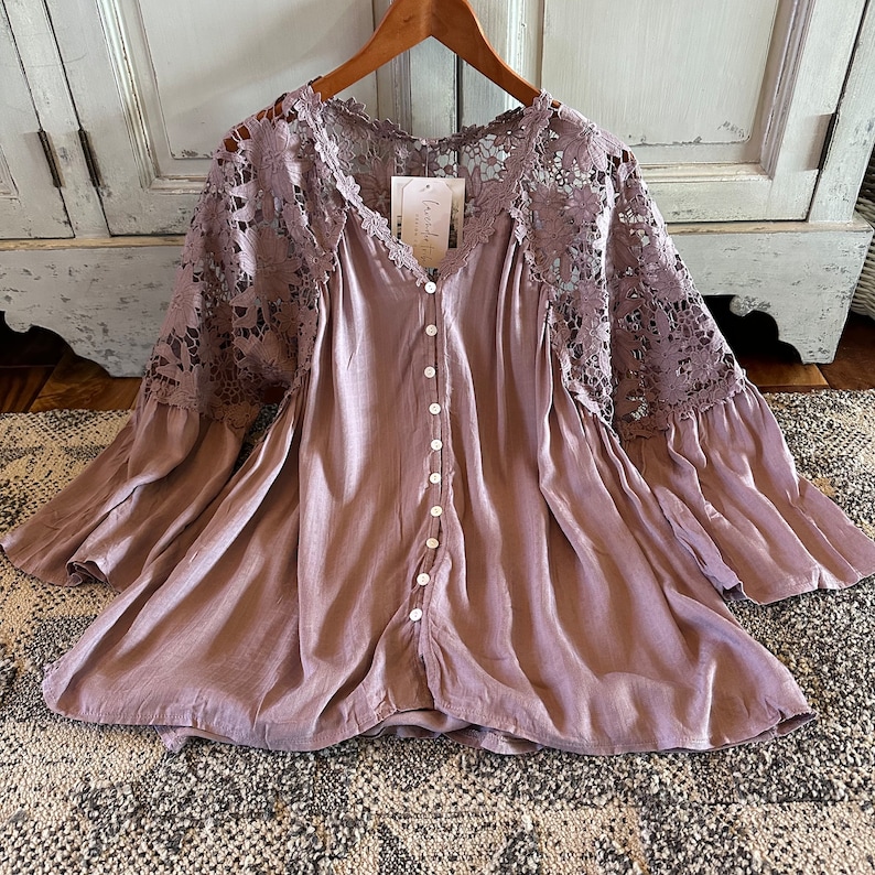 May include: A mauve button-up blouse with a lace overlay on the sleeves. The blouse has a relaxed fit and a slightly flared hem.