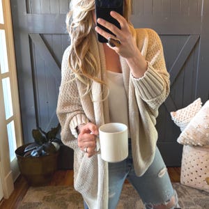 May include: A beige open-front knit cardigan with a textured stitch pattern and ribbed cuffs, worn over a white top and light blue distressed denim jeans, while holding a plain white ceramic mug in one hand.