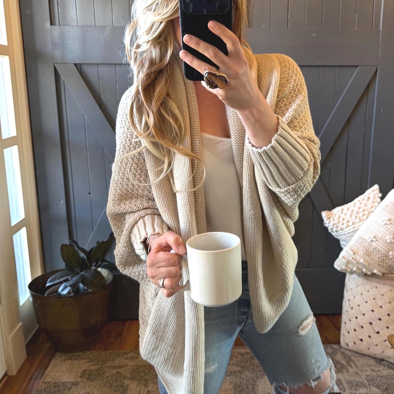 May include: A beige open-front knit cardigan with a textured stitch pattern and ribbed cuffs, worn over a white top and light blue distressed denim jeans, while holding a plain white ceramic mug in one hand.