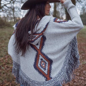 May include: A gray hooded poncho with a geometric pattern in shades of orange and navy blue. The poncho has a fringed hem and is paired with a brown cowboy hat. The outfit is completed with blue jeans.