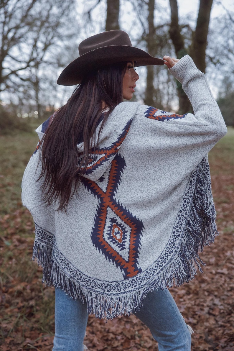 May include: A gray hooded poncho with a geometric pattern in shades of orange and navy blue. The poncho has a fringed hem and is paired with a brown cowboy hat. The outfit is completed with blue jeans.