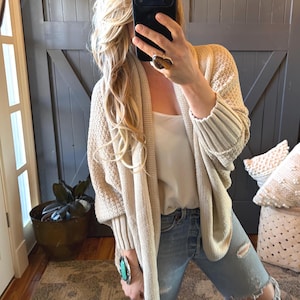 May include: A beige knit cardigan with a shawl collar and ribbed cuffs. The cardigan is worn over a white tank top and paired with distressed blue jeans. A large ring and bracelet are visible.