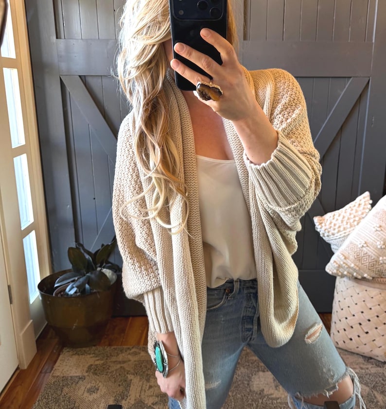 May include: A beige knit cardigan with a shawl collar and ribbed cuffs. The cardigan is worn over a white tank top and paired with distressed blue jeans. A large ring and bracelet are visible.
