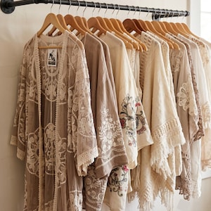 May include: A row of beige and cream-colored kimonos and cardigans hanging on a black industrial pipe rack. The garments feature intricate lace, embroidery, and fringe details. Each piece is displayed on a wooden hanger against a plain wall.
